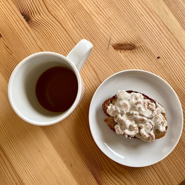 Eine Scheibe Brot mit Nordseekrabbensalat auf einem Holztisch. Daneben eine Tasse Kaffee.