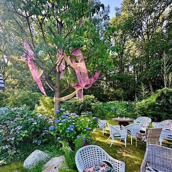 Morgensonne im Garten, ein Baum mit bunten Schleiern und weissen Korbstühlen.